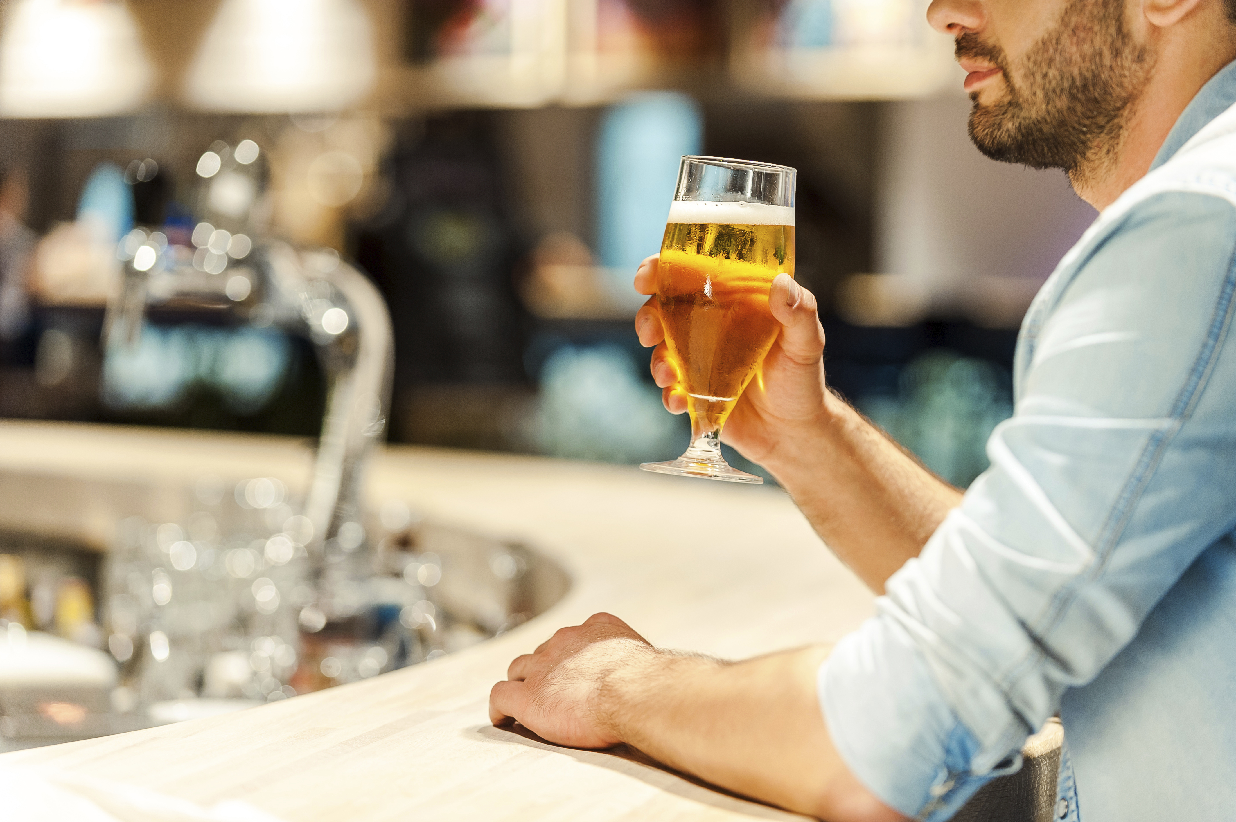 Man holding a glass of beer while sitting at a bar counter with a blurred background of bottles and taps.