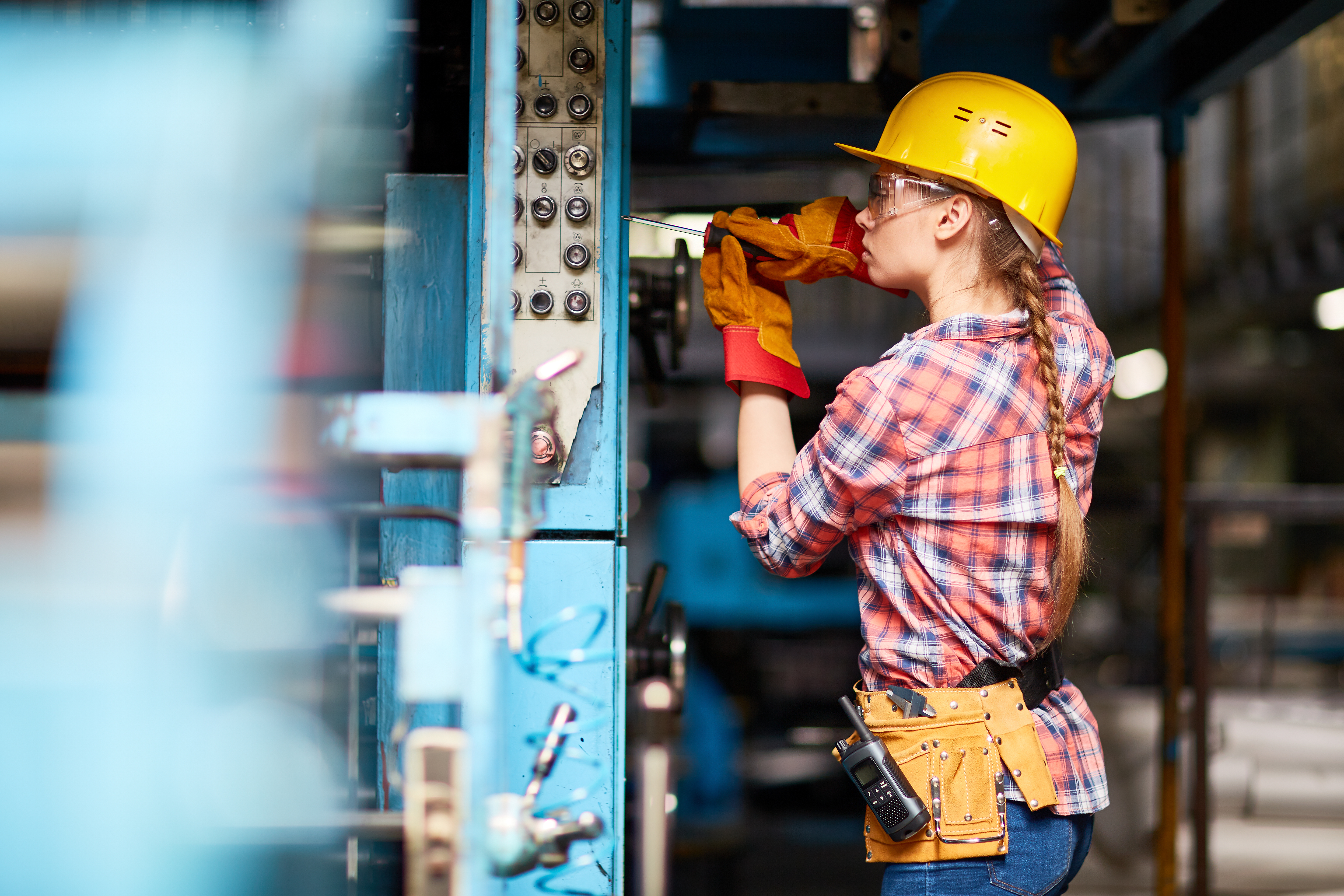 Female factory worker wearing a yellow hard hat and plaid shirt operating machinery in an industrial setting.