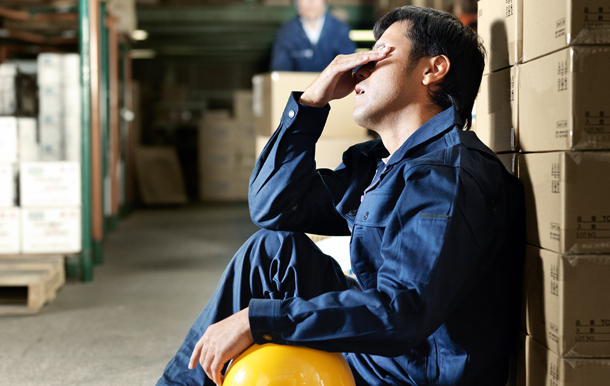Male warehouse worker in blue uniform sitting on the floor, resting against stacked boxes, holding a yellow hard hat and wiping sweat from his forehead.