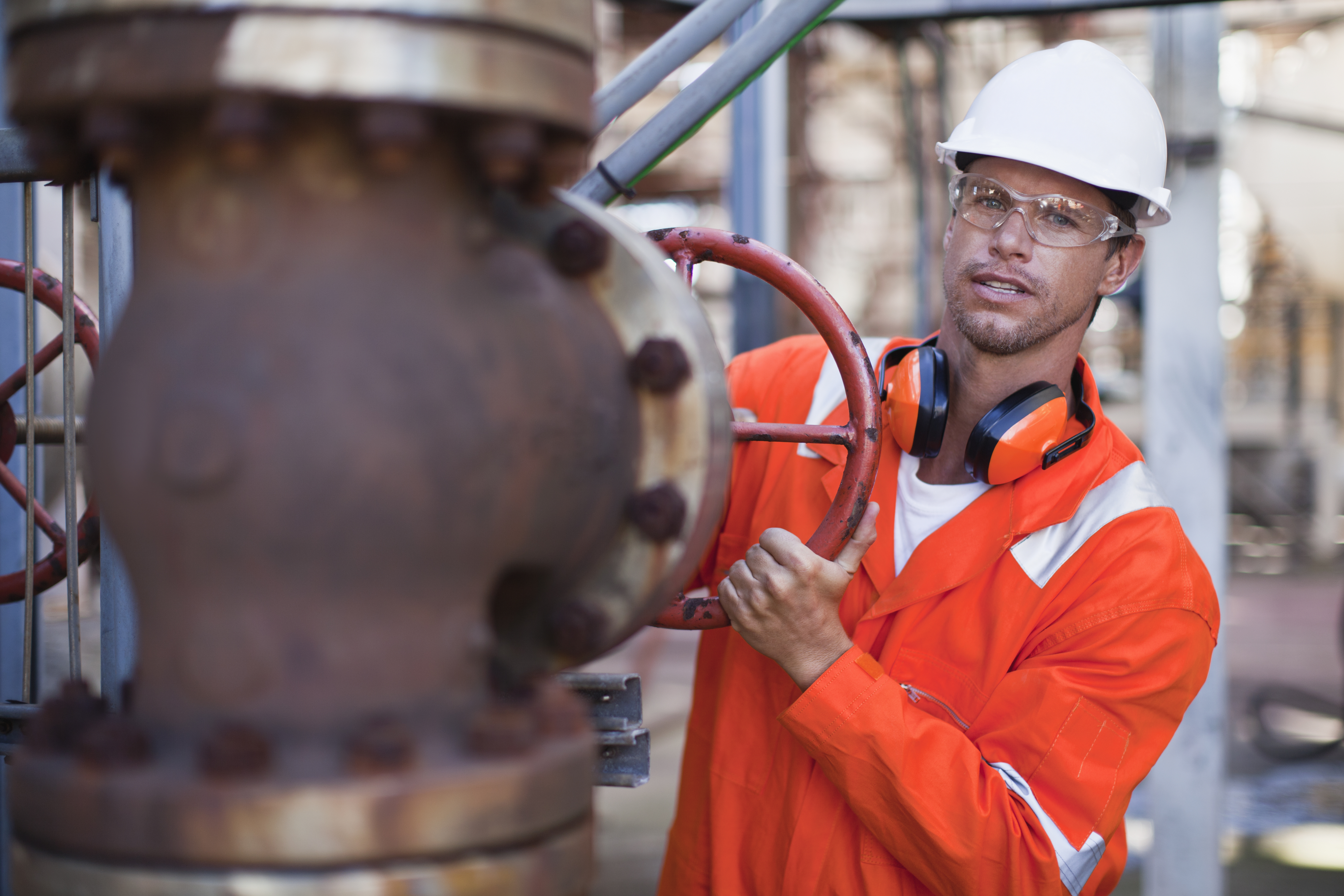 Male industrial worker in orange safety gear and white helmet turning a large red valve on a rusted pipe system outdoors.