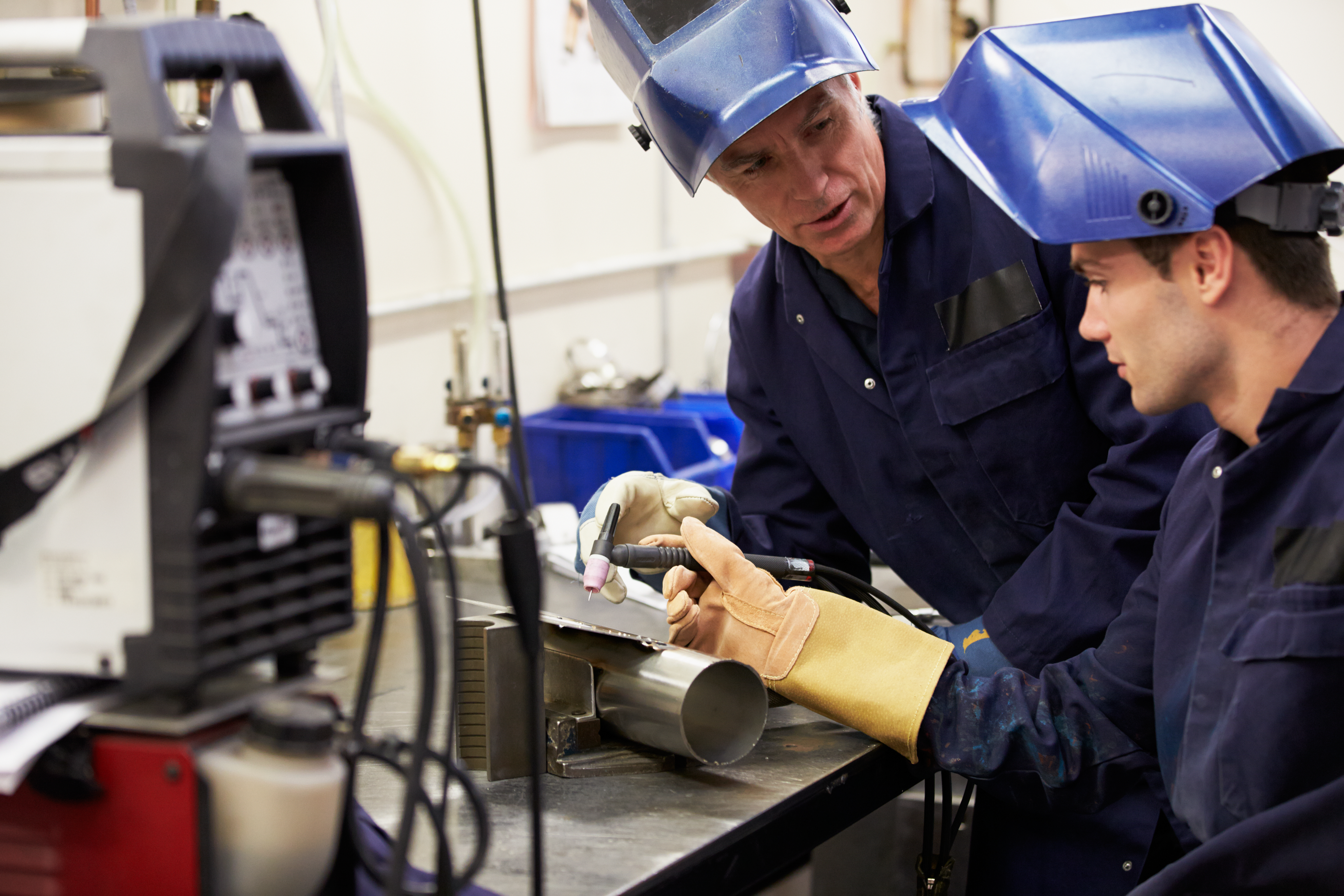 Two workers in protective gear welding a metal pipe in an industrial workshop setting.