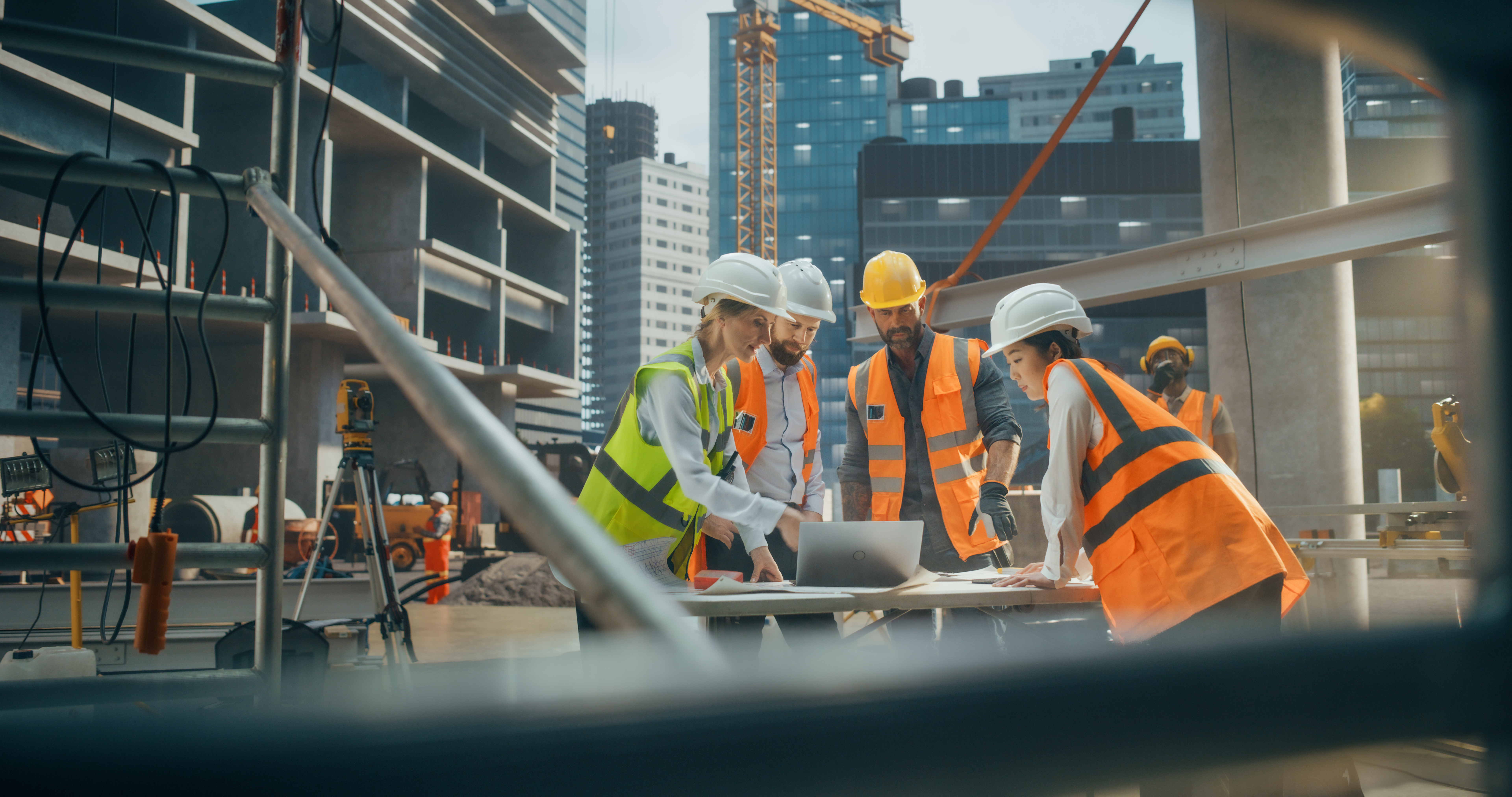 Construction workers in safety gear reviewing blueprints on an urban building site with cranes and skyscrapers in the background.