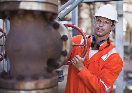 Male industrial worker in orange safety gear and white helmet turning a large red valve on a rusted pipe system outdoors.