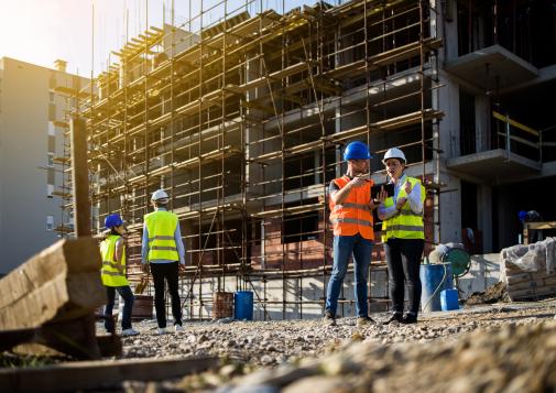 Construction workers in safety gear discussing plans at a building site with scaffolding in the background during sunset.