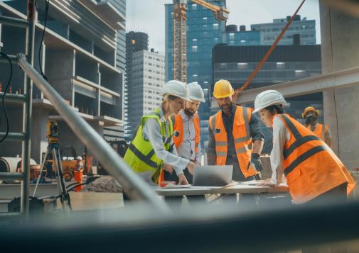 Construction workers in safety gear reviewing blueprints on an urban building site with cranes and skyscrapers in the background.