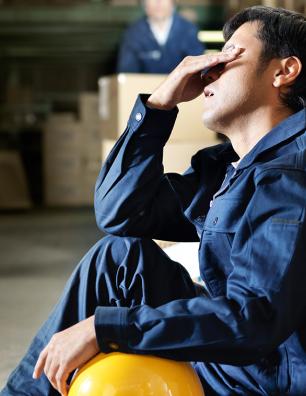 Male warehouse worker in blue uniform sitting on the floor, resting against stacked boxes, holding a yellow hard hat and wiping sweat from his forehead.