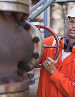 Male industrial worker in orange safety gear and white helmet turning a large red valve on a rusted pipe system outdoors.