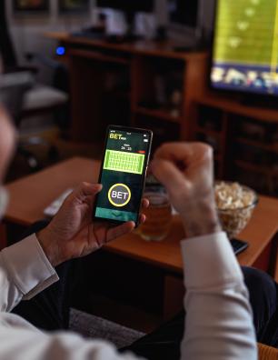 Man holding smartphone with a betting app open while watching a football game on TV in a cozy living room.
